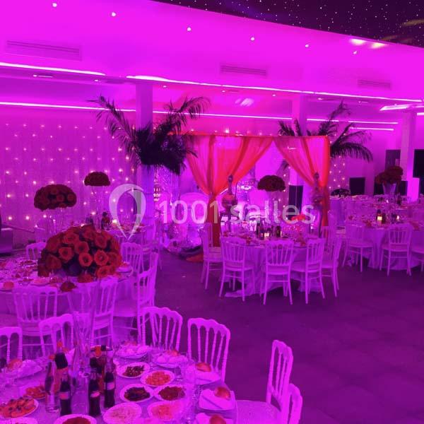 Location salle Le Raincy (Seine-Saint-Denis) - Le Chalet des Pins #8 Salle de réception décorée avec des tables dressées, éclairage rose vif et rideaux ornés de fleurs rouges.