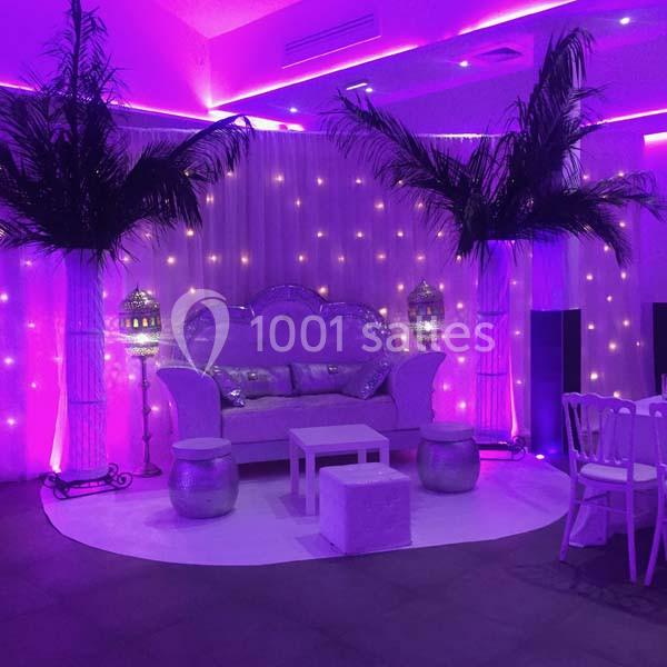 Location salle Le Raincy (Seine-Saint-Denis) - Le Chalet des Pins #11 Salon décoré avec des lumières violettes, des palmiers artificiels, un canapé argenté et des lanternes orientales.
