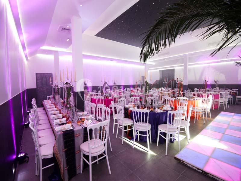 Location salle Le Raincy (Seine-Saint-Denis) - Le Chalet des Pins #2 Salle de réception décorée avec des tables dressées, des chaises blanches et un éclairage violet tamisé.