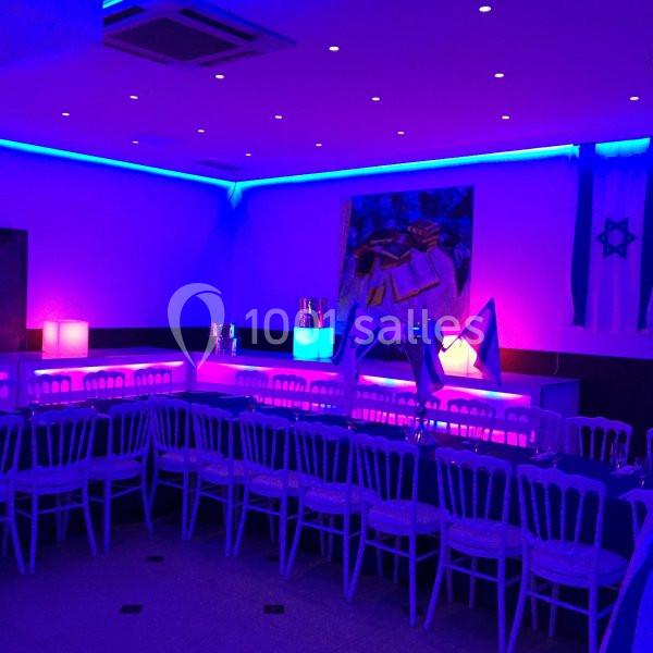 Location salle Le Raincy (Seine-Saint-Denis) - Le Chalet des Pins #21 Salle décorée avec des lumières bleues et violettes, tables alignées avec chaises blanches, drapeaux visibles en arrière…