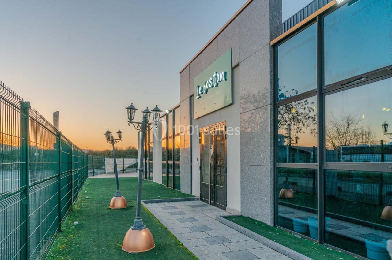 Façade d'un bâtiment moderne avec grandes baies vitrées, éclairage extérieur et clôture métallique au coucher du soleil.