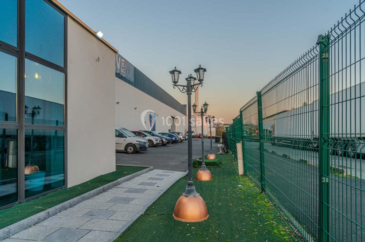 Allée bordée de lampadaires et clôturée, menant à un bâtiment industriel avec des voitures stationnées.