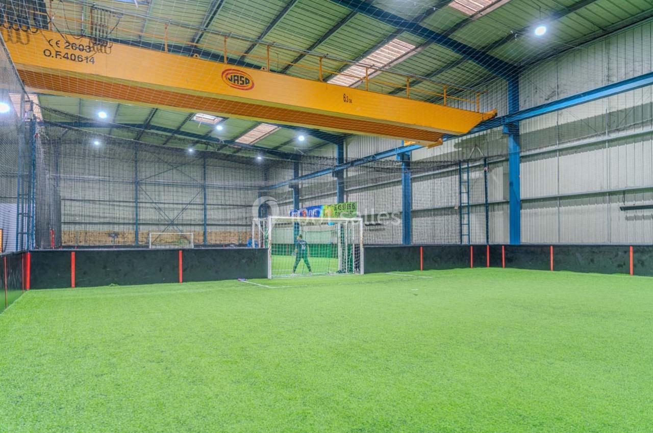Terrain de football en salle avec gazon synthétique, filet de protection et structure métallique visible en arrière-plan.