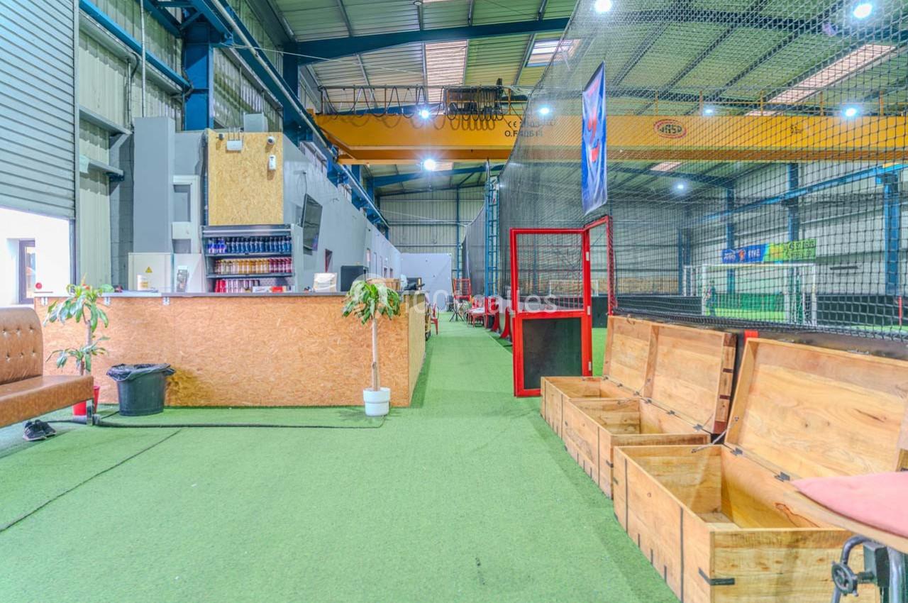 Espace intérieur avec comptoir en bois, bancs ouverts, sol en gazon synthétique et terrain de sport grillagé au fond.