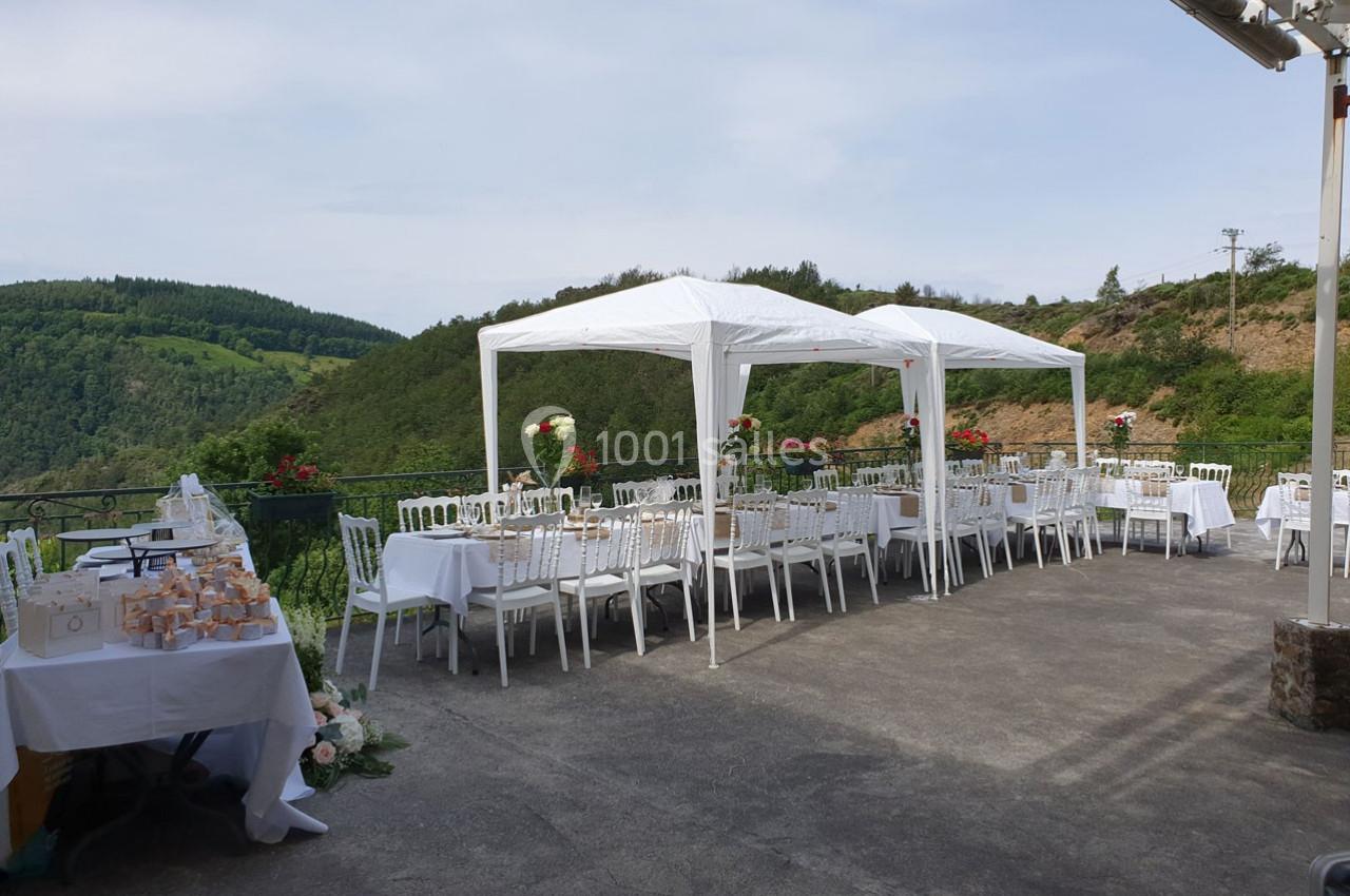 Tables et chaises blanches disposées sous des tonnelles sur une terrasse avec vue sur des collines verdoyantes.
