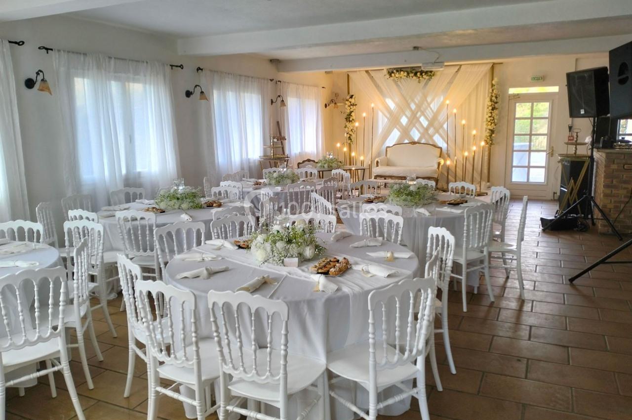 Salle de réception décorée pour un événement, avec des tables rondes dressées, chaises blanches et éclairage chaleureux.
