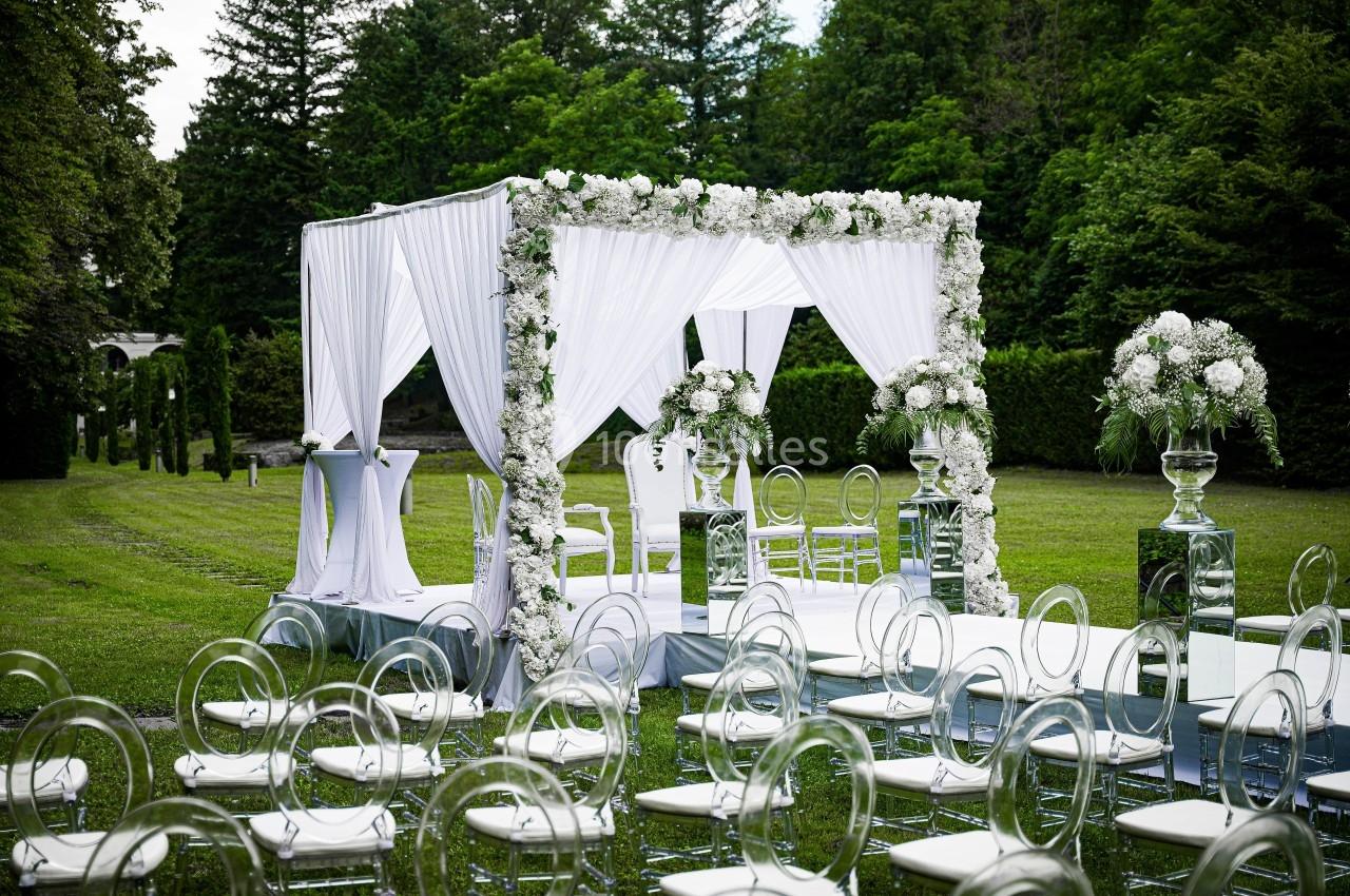 Structure de cérémonie de mariage en plein air avec arche décorée de fleurs blanches et chaises transparentes sur pelouse…
