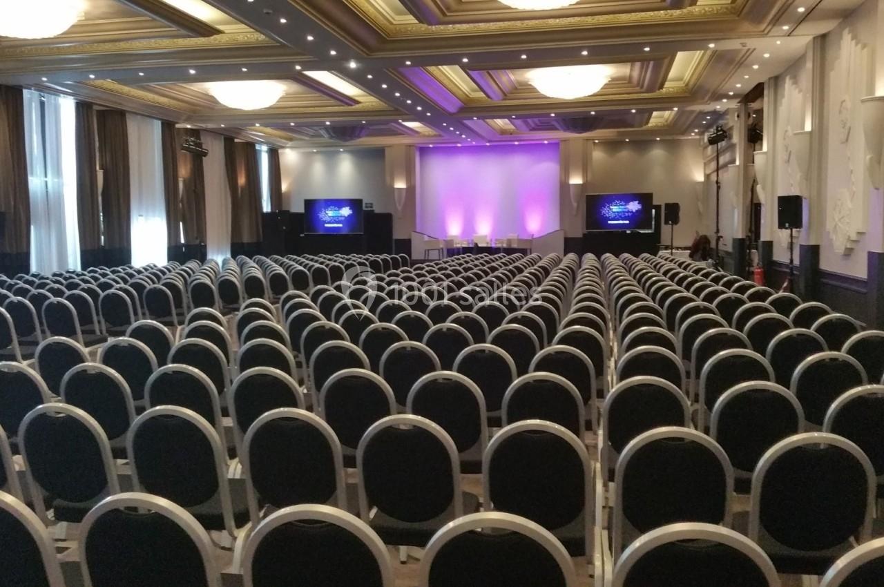 Salle de conférence avec rangées de chaises noires alignées face à une scène éclairée de lumières violettes.