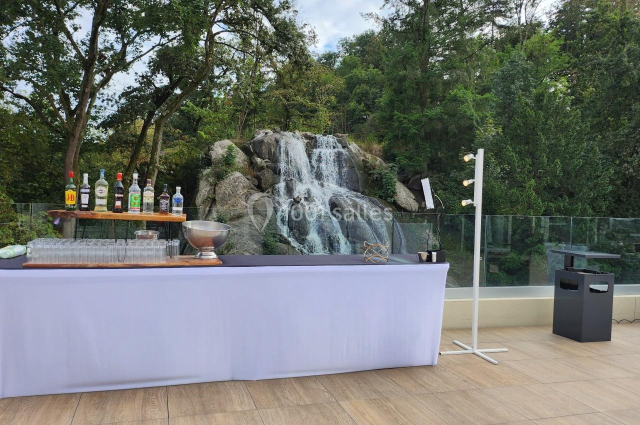 Table de bar extérieure avec bouteilles, verres et accessoires, devant une cascade entourée de végétation.