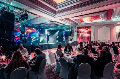Des invités dînent autour de tables rondes tandis qu'un groupe joue sur une scène illuminée dans une salle de réception.