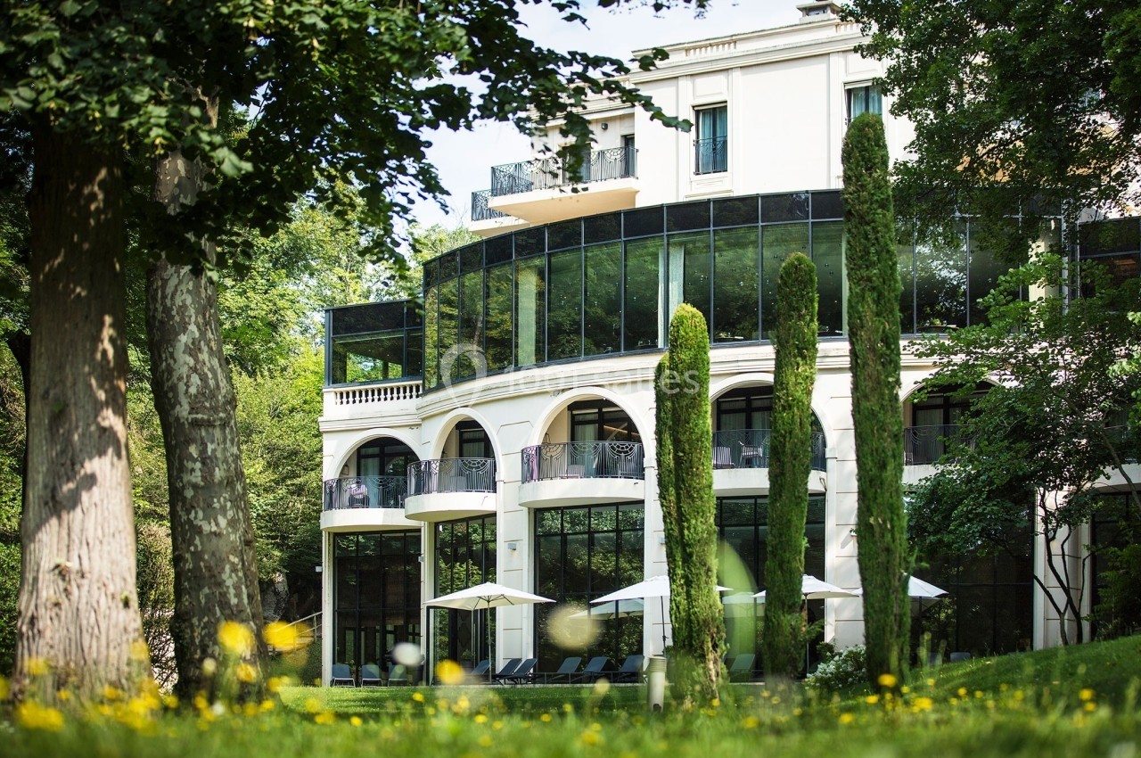 Façade d'un bâtiment moderne entouré d'arbres, avec des baies vitrées et une terrasse équipée de parasols.