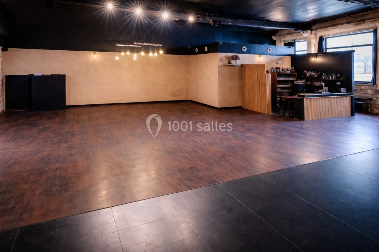Salle vide avec parquet, éclairage suspendu, bar en bois à droite et murs clairs dans un espace industriel.