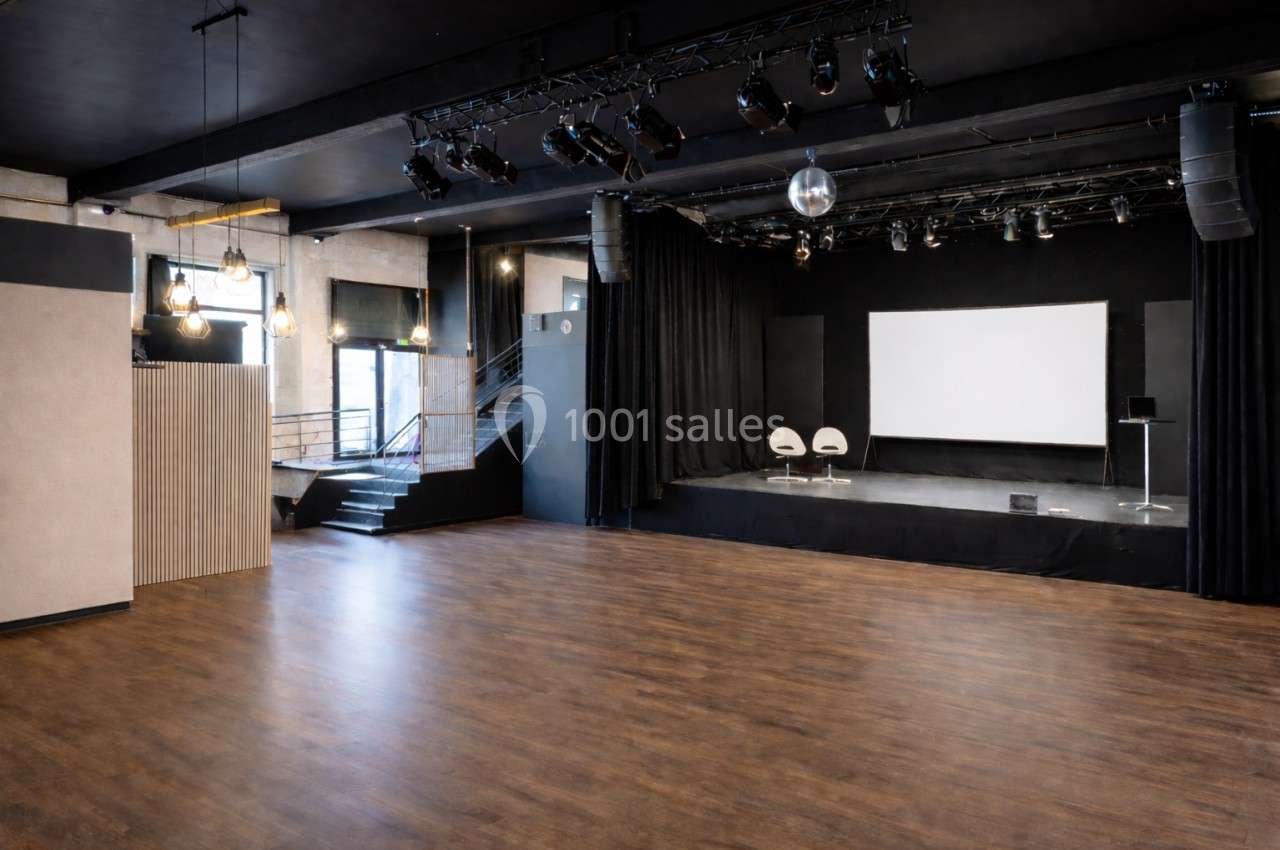 Salle polyvalente avec parquet, scène équipée de rideaux noirs, écran blanc et projecteurs suspendus.