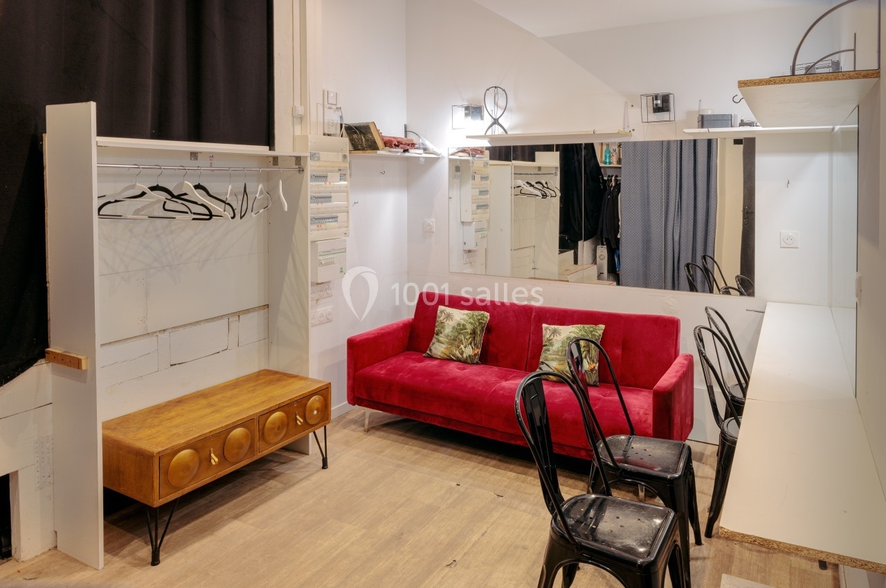 Loge avec canapé rouge, miroir mural, chaises noires, table en bois et portants avec cintres dans un espace lumineux.