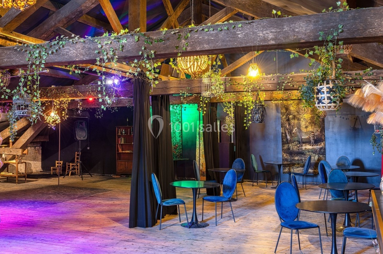 Salle avec poutres apparentes, décorée de plantes suspendues, éclairage coloré et mobilier composé de tables et chaises…