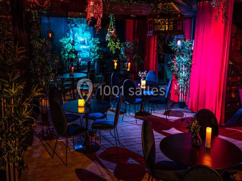 Salle sombre avec tables rondes, chaises noires, bougies allumées et rideaux rouges, ambiance intime et feutrée.