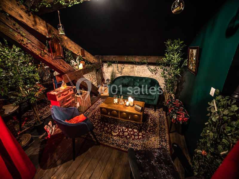 Salon chaleureux sous combles avec canapé vert, tapis persan, meubles en bois, plantes et éclairage tamisé.