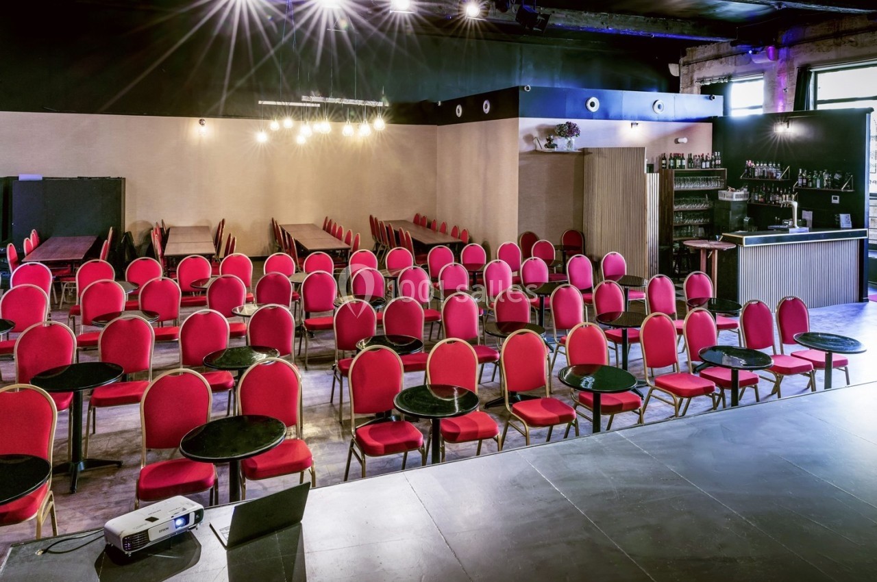 Salle de conférence avec rangées de chaises rouges et petites tables rondes, vue depuis une scène.