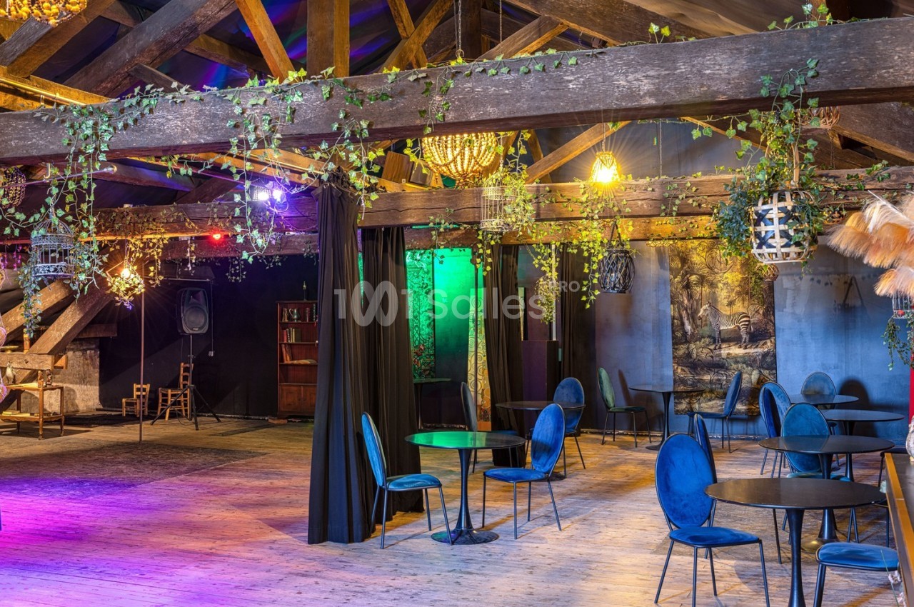 Salle avec poutres apparentes, décorée de plantes suspendues, éclairage coloré et mobilier composé de tables et chaises…