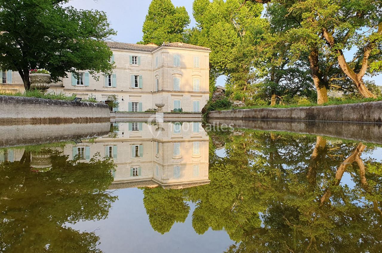 Reflet d'un bâtiment ancien en pierre et d'arbres dans un bassin calme, entouré de verdure.
