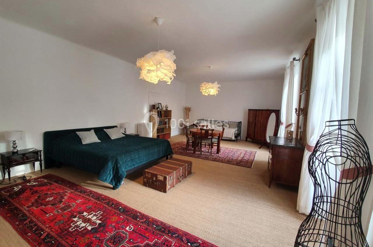 Chambre spacieuse avec lit double, table à manger, tapis rouges, meubles en bois et grandes fenêtres avec rideaux blancs.
