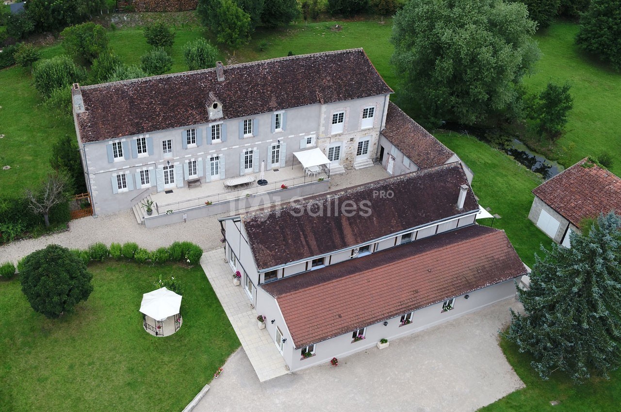 Vue aérienne d'un grand bâtiment ancien avec toiture en tuiles, entouré de jardins et d'arbres verdoyants.