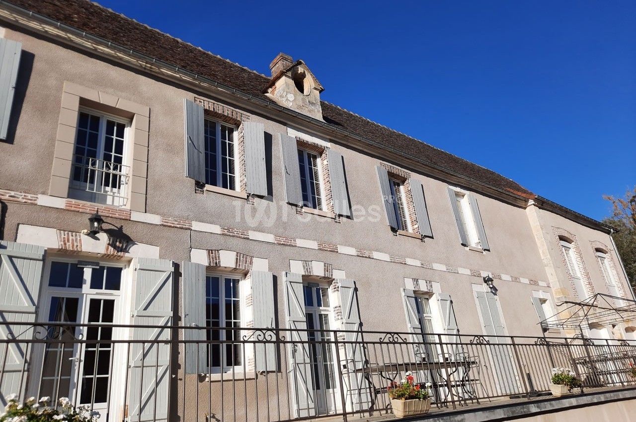 Façade d'une maison traditionnelle avec volets clairs, terrasse et ciel bleu en arrière-plan.