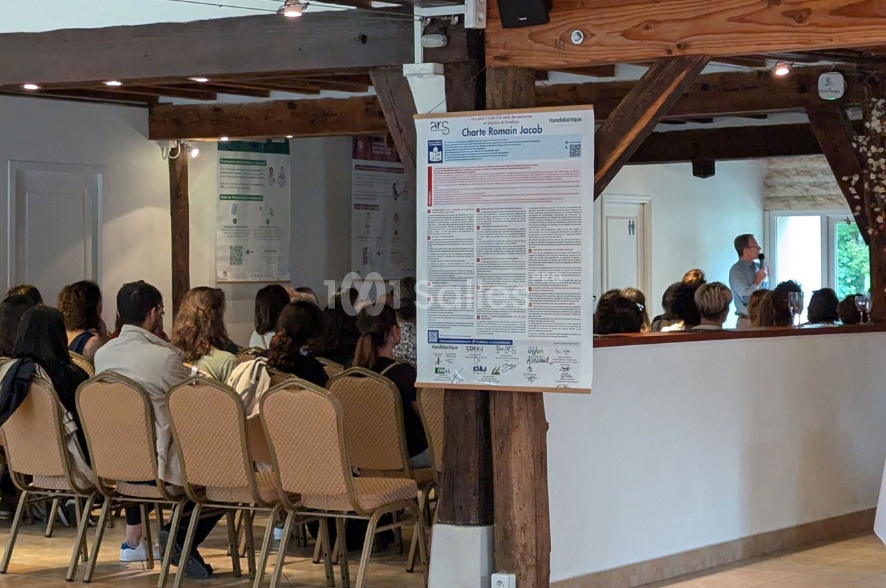 Un groupe de personnes assises dans une salle, écoutant un intervenant debout près d'un mur en bois.