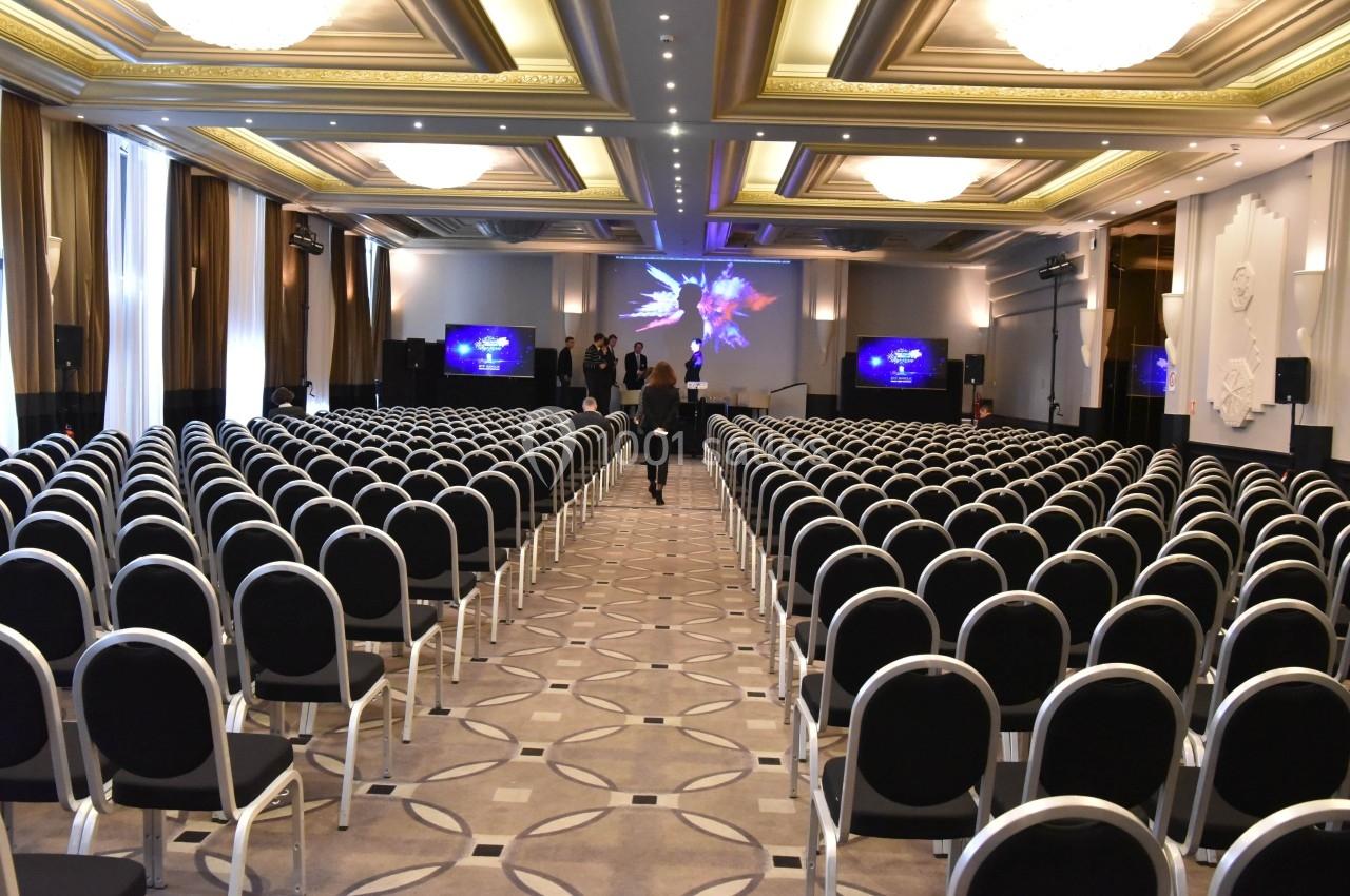 Location salle La Tour-de-Salvagny (Rhône) - Le Pasino Grand et l'Hôtel & Spa Le Pavillon #2 Salle de conférence vide avec des rangées de chaises noires, un écran et une scène éclairée en arrière-plan.