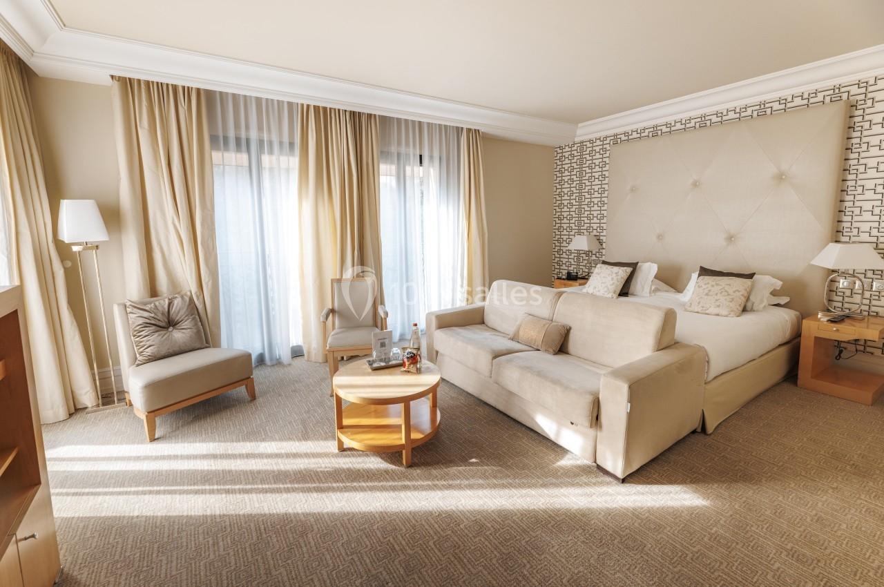 Location salle La Tour-de-Salvagny (Rhône) - Le Pasino Grand et l'Hôtel & Spa Le Pavillon #9 Chambre lumineuse avec lit double, canapé, fauteuils, table basse et grandes fenêtres avec rideaux beiges.