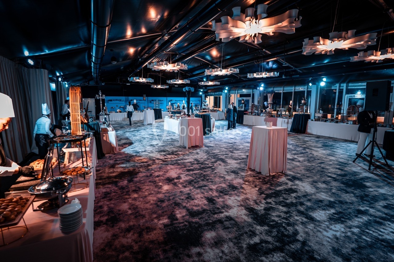 Salle de réception éclairée avec des buffets dressés, des tables hautes et des chefs en train de préparer des plats.