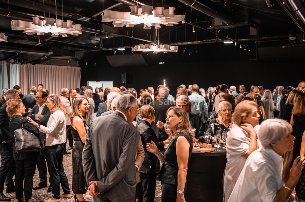 Groupe de personnes échangeant dans une salle de réception éclairée, avec des tables hautes et des rideaux en arrière-plan.