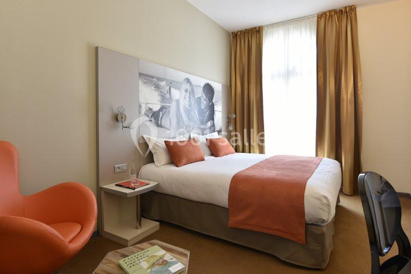 Chambre d'hôtel lumineuse avec un lit double, des coussins orange, un fauteuil assorti et une grande fenêtre avec rideaux…