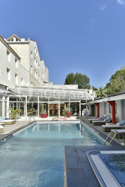 Piscine extérieure entourée de transats et d'un bâtiment blanc avec une verrière, sous un ciel bleu clair.