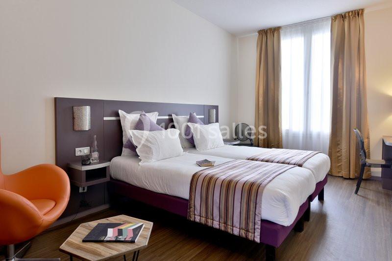 Chambre d'hôtel lumineuse avec deux lits simples, des coussins violets, un fauteuil orange et un bureau près de la fenêtre.