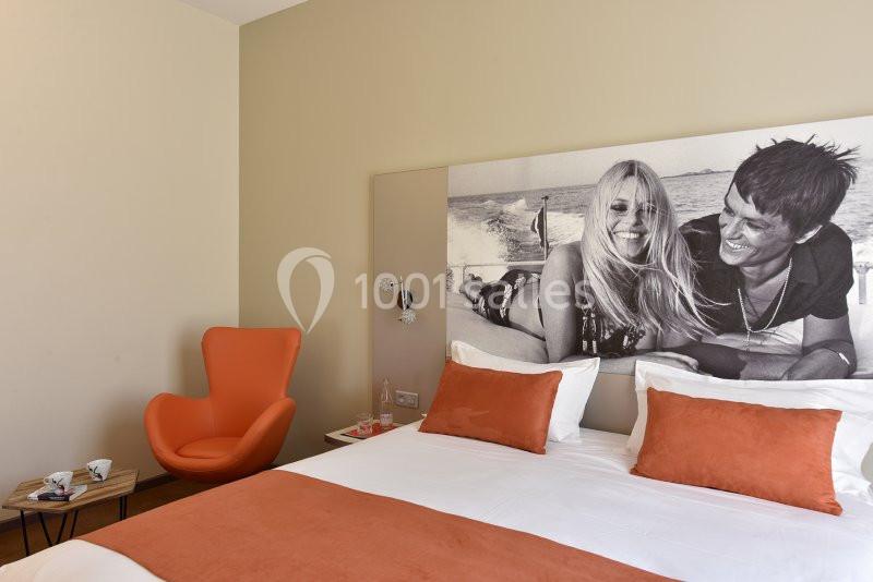 Chambre d'hôtel avec lit double, coussins orange, fauteuil assorti et grande photo en noir et blanc au mur.