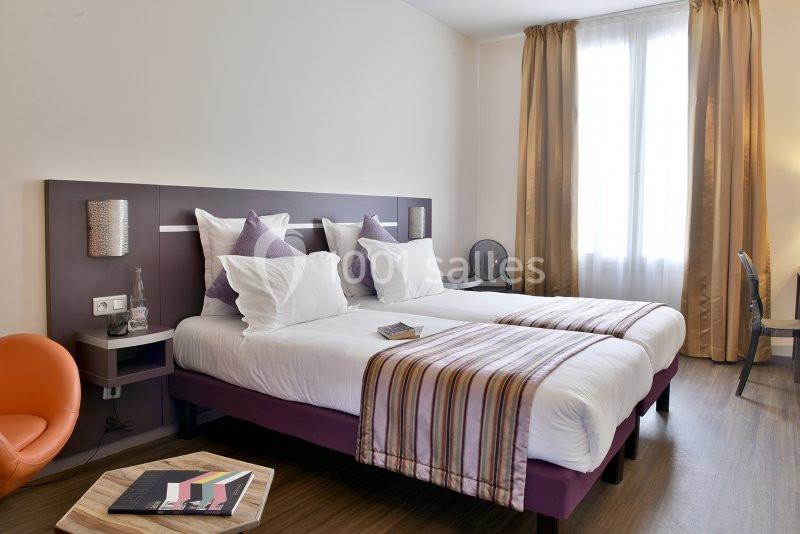 Chambre lumineuse avec deux lits simples, draps blancs, coussins violets, bureau, fauteuil orange et grande fenêtre avec…