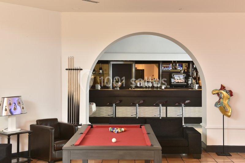 Salle avec une table de billard rouge, un bar en arrière-plan et des fauteuils en cuir noir.