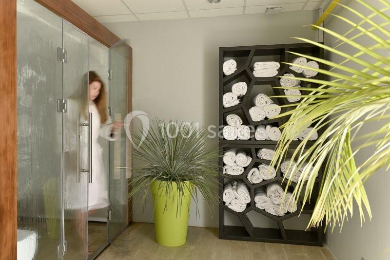 Une femme en peignoir sort d'un sauna vitré, près d'une étagère remplie de serviettes roulées et de plantes vertes.