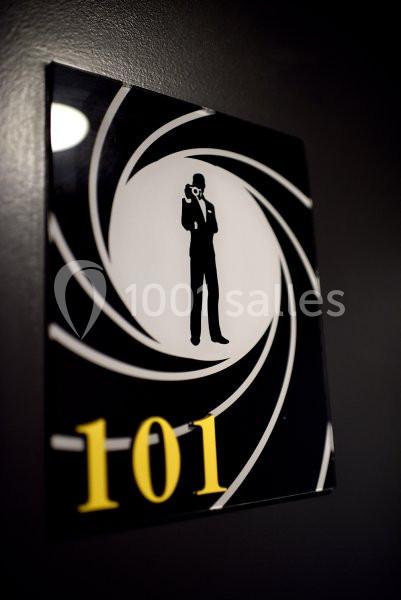 Plaque noire avec le chiffre ’101’ en jaune et une silhouette d'espion dans un viseur stylisé.