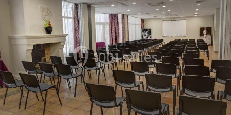 Salle de conférence avec rangées de chaises noires, écran blanc au fond et cheminée décorée sur la gauche.
