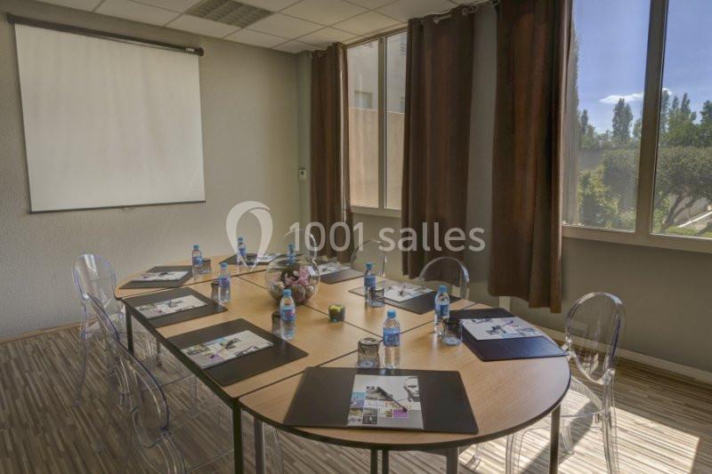 Salle de réunion lumineuse avec table ovale, chaises transparentes, documents, bouteilles d'eau et écran de projection.