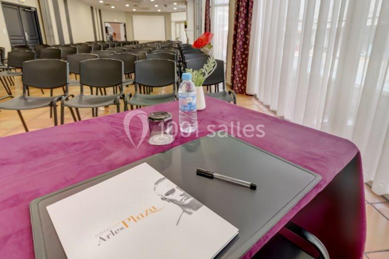 Table recouverte d'une nappe violette avec un carnet, un stylo, une bouteille d'eau et une rose, face à des chaises alignées.
