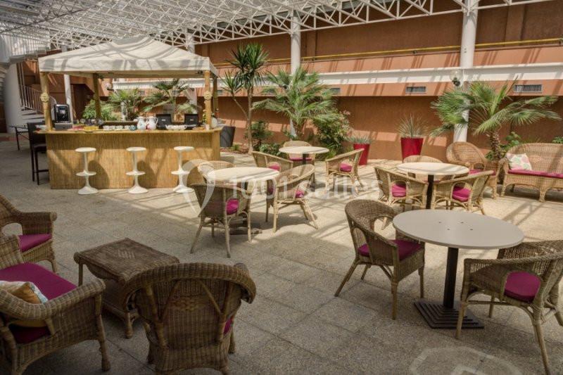 Terrasse couverte avec mobilier en rotin, tables rondes, plantes et un comptoir de bar en arrière-plan.