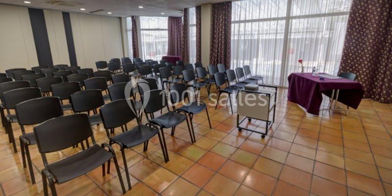Salle de conférence avec rangées de chaises noires, une table violette et de grandes fenêtres avec rideaux.