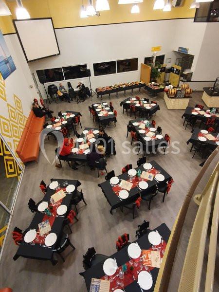 Salle de réception vue de haut, tables dressées avec nappes noires et assiettes blanches, quelques personnes assises.
