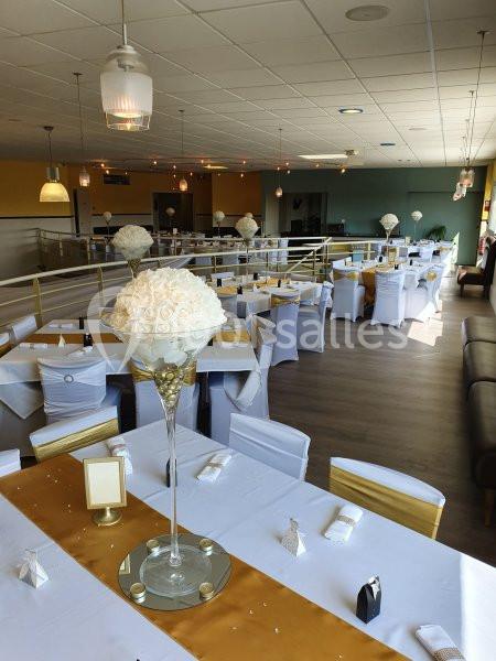 Salle de réception décorée avec des tables dressées, nappes blanches, chemins de table dorés et centres de table fleuris.