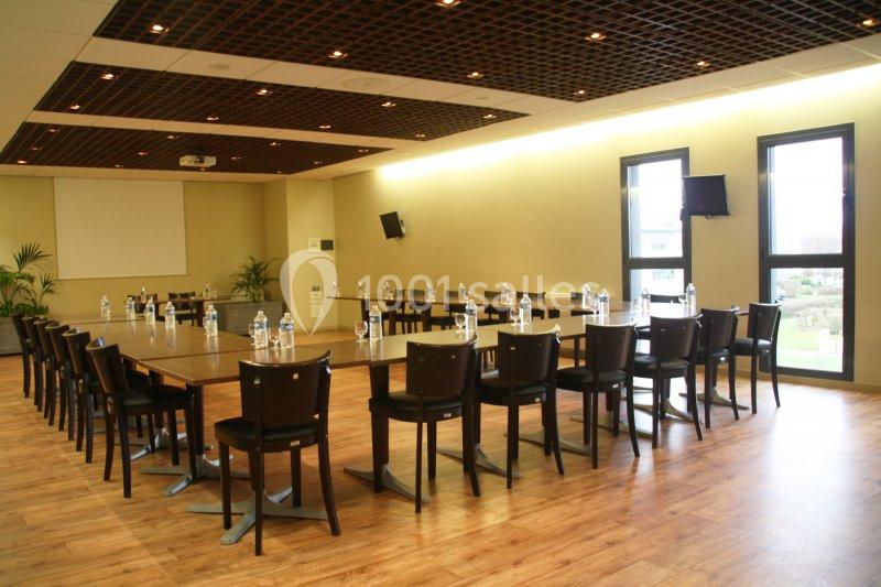Salle de réunion lumineuse avec tables disposées en U, chaises noires, bouteilles d'eau et écran de projection.