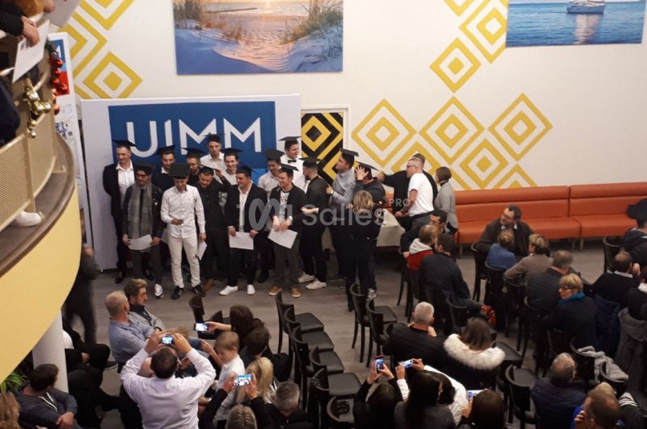 Un groupe de jeunes hommes posant devant un panneau, entourés d’un public assis dans une salle décorée.