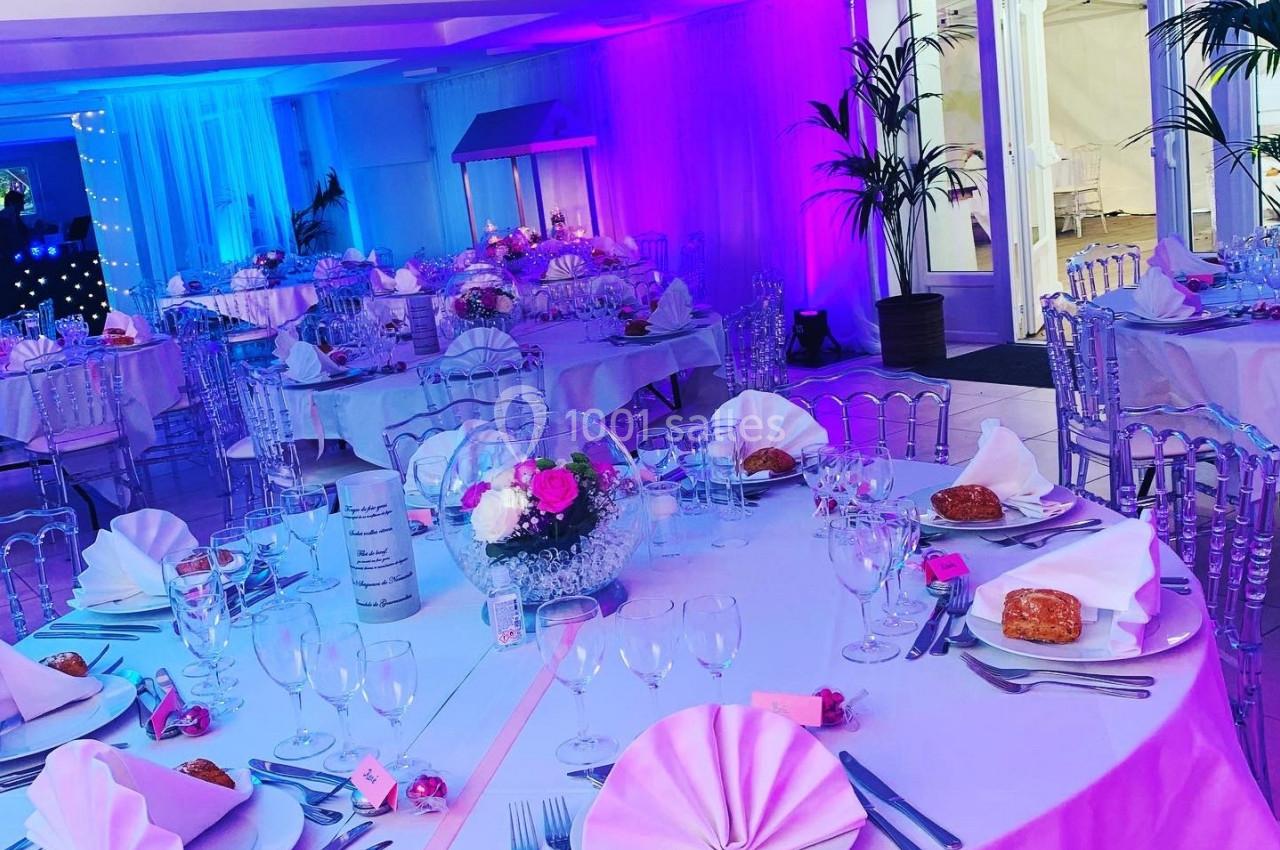 Salle de réception décorée avec des tables rondes, nappes blanches, serviettes roses et éclairage coloré.